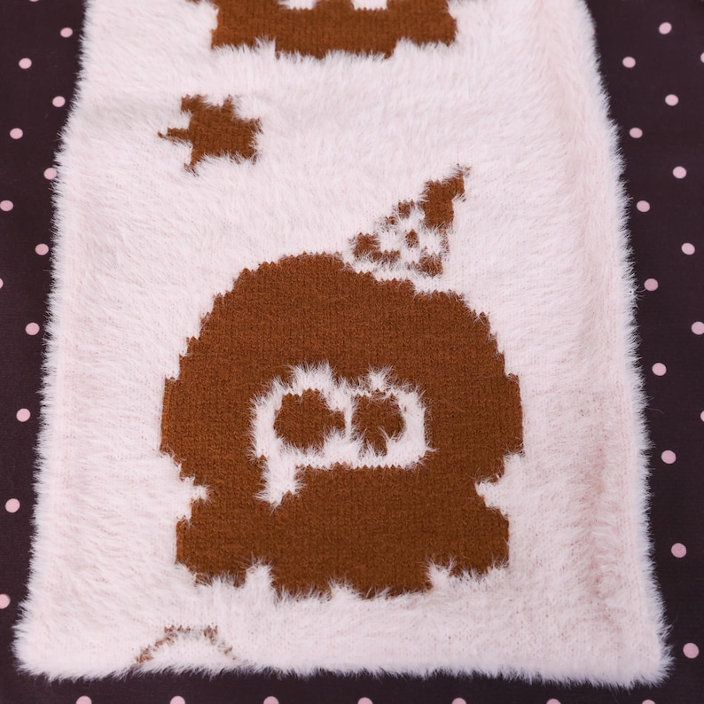 May include: A close-up of a white and brown fuzzy textile with a repeating pattern. The pattern features a brown ghost-like figure with a star and other shapes. The background is a dark brown with white polka dots.