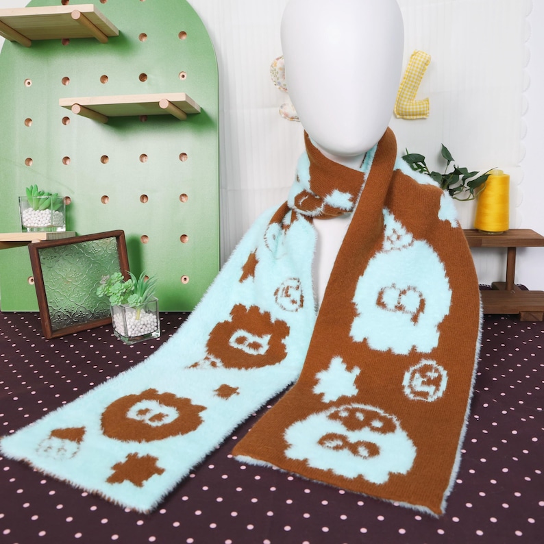 May include: A fluffy scarf with a light blue and brown pattern. The scarf features repeating cartoon character designs. The scarf is displayed on a mannequin head. The scarf is made of a soft, fuzzy material.