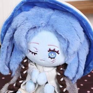May include: A plush doll with a light blue face, blue hair, and a blue hooded hat. The doll has embroidered facial features, including closed eyes and a single open eye. It wears a white outfit with brown braids.