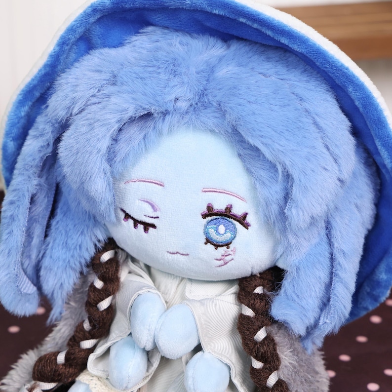 May include: A plush doll with a light blue face, blue hair, and a blue hooded hat. The doll has embroidered facial features, including closed eyes and a single open eye. It wears a white outfit with brown braids.