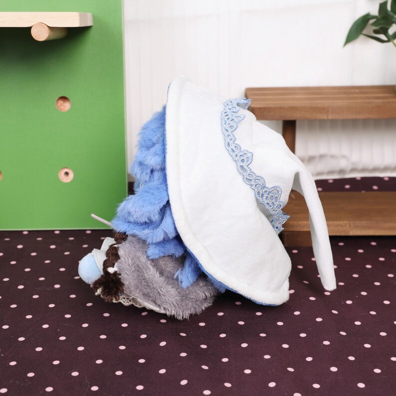 May include: A plush toy featuring a white bonnet with blue lace trim and long, floppy ears. The toy has blue and gray fur, and a small, light blue pom-pom. The toy is lying on a polka-dot surface.