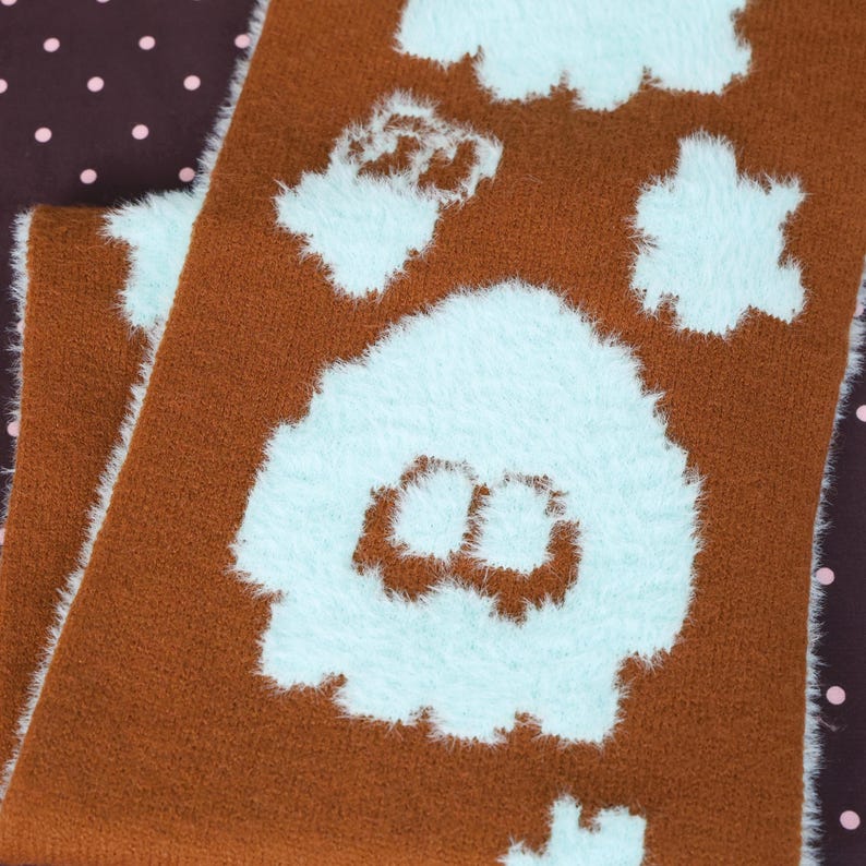 May include: A brown scarf with a repeating pattern of fluffy, light blue cloud-like shapes. The scarf has a soft, textured appearance and is folded over itself. The background is a dark brown fabric with pink polka dots.