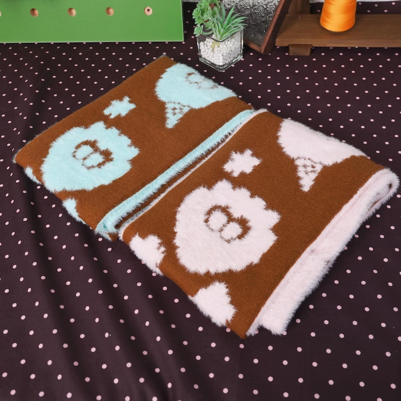 May include: Two folded brown blankets with a repeating pattern of a bear's face in white and light blue. The blankets have a soft, plush texture and are displayed on a brown polka dot surface.