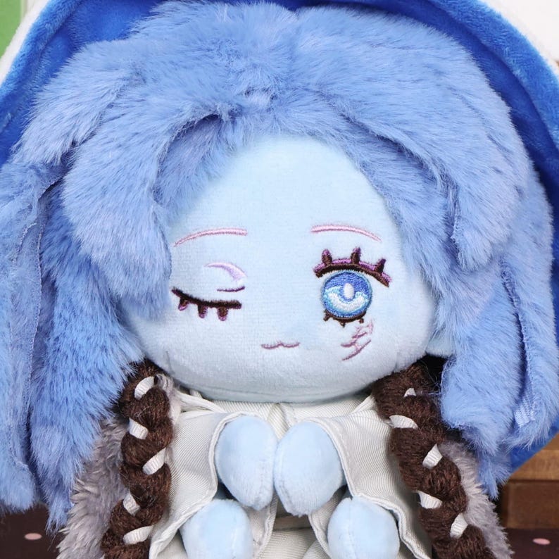 May include: A plush doll with a light blue face, winking eye, and embroidered details. The doll has blue hair, a blue hood, and a white and gray outfit. Brown braids frame the face. The doll is a soft toy.