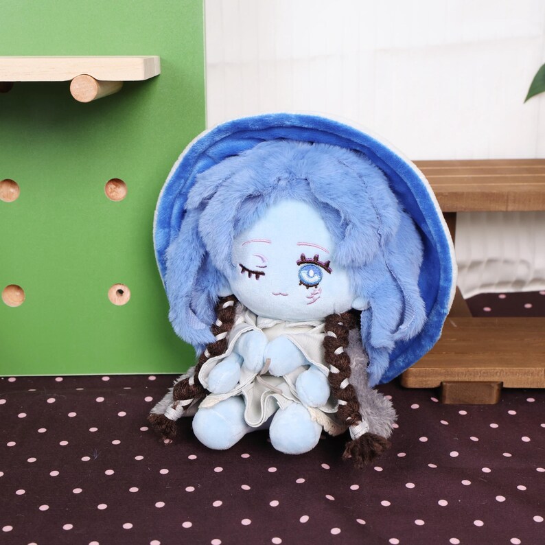 May include: A plush doll with a blue face, hair, and outfit. The doll has closed eyes, a small smile, and brown braids. It is sitting on a black and white polka dot surface.