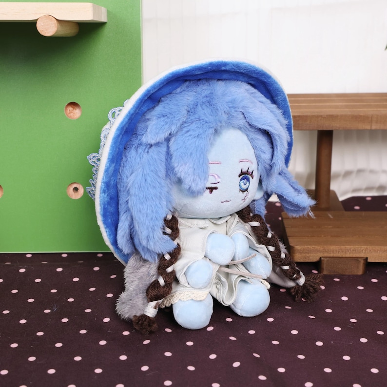 May include: A plush doll with blue fur hair and a blue bonnet. The doll has a light blue face with a small smile and a white dress. The doll has brown braids and is sitting on a polka dot surface.