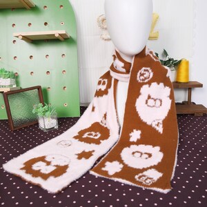 May include: A brown and pink scarf with a repeating pattern of cartoon bear faces and floral shapes. The scarf is made of a soft, fuzzy material and is draped over a mannequin. The scarf is a winter accessory.