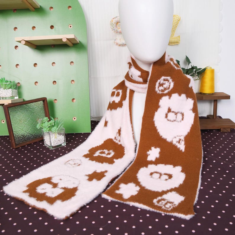May include: A brown and pink scarf with a repeating pattern of cartoon bear faces and floral shapes. The scarf is made of a soft, fuzzy material and is draped over a mannequin. The scarf is a winter accessory.