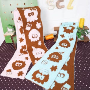 May include: Three fluffy scarves in pink, brown, and blue. Each scarf features a repeating pattern of cartoon-like characters and star shapes. The scarves are displayed on a polka-dot surface, suggesting a cozy, winter theme.
