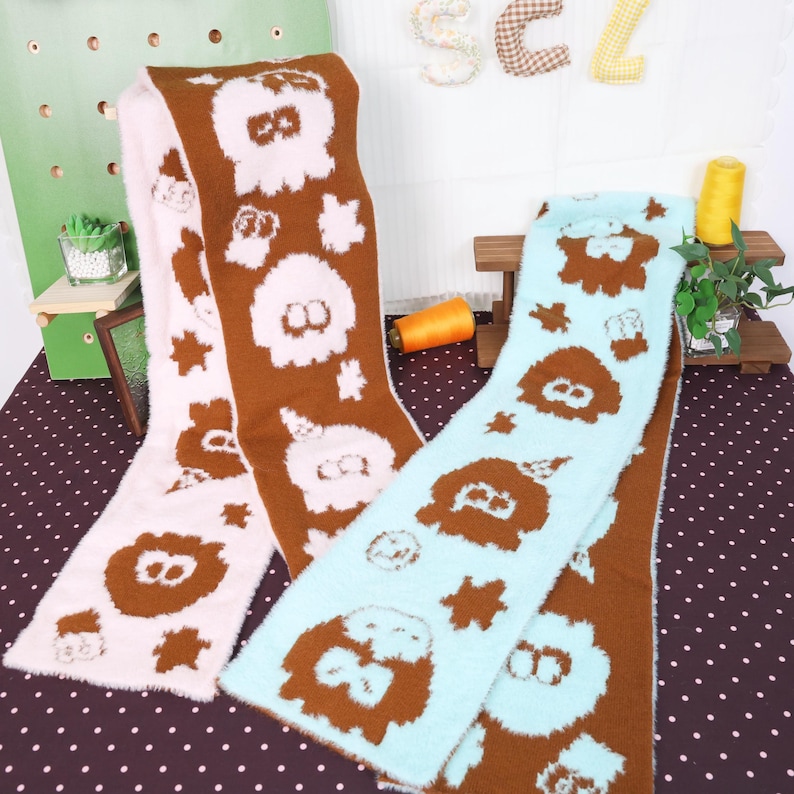 May include: Three fluffy scarves in pink, brown, and blue. Each scarf features a repeating pattern of cartoon-like characters and star shapes. The scarves are displayed on a polka-dot surface, suggesting a cozy, winter theme.