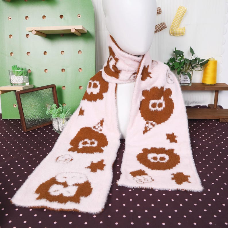 May include: A light pink scarf with brown bear-like character designs. The scarf is displayed on a mannequin and has a soft, fuzzy texture. The bear designs are repeated along the length of the scarf.