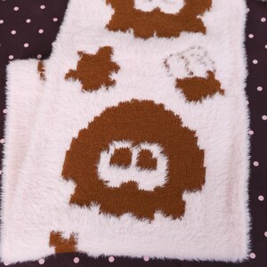 May include: A white and brown fuzzy textile with a repeating pattern of cartoon ghost-like figures. The figures are brown and have a simple, rounded design. The background is a dark brown fabric with small white polka dots.