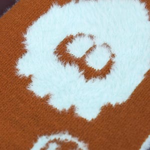 May include: Close-up of a brown textile with a light blue, fluffy, abstract design. The design appears to be a stylized letter or shape. The textile has a soft, textured appearance.