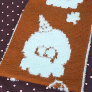May include: Close-up of a brown and white plush textile with a cartoon ghost design. The ghost is white with a light blue outline and a simple face. The background is a dark brown fabric with pink polka dots.
