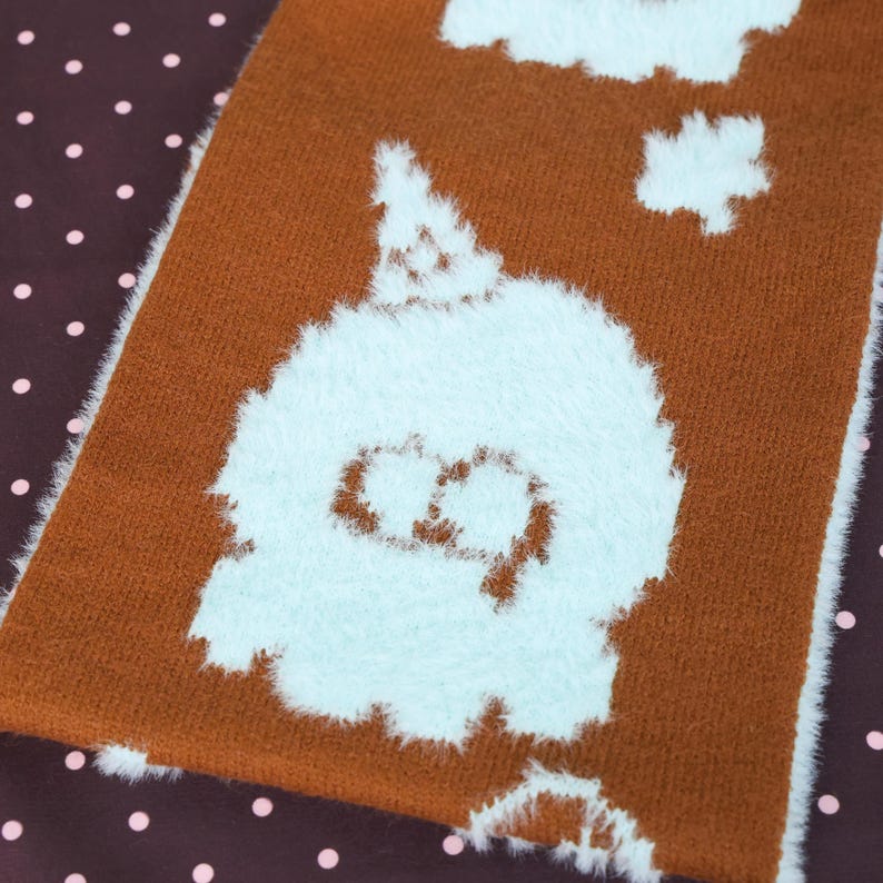 May include: Close-up of a brown and white plush textile with a cartoon ghost design. The ghost is white with a light blue outline and a simple face. The background is a dark brown fabric with pink polka dots.