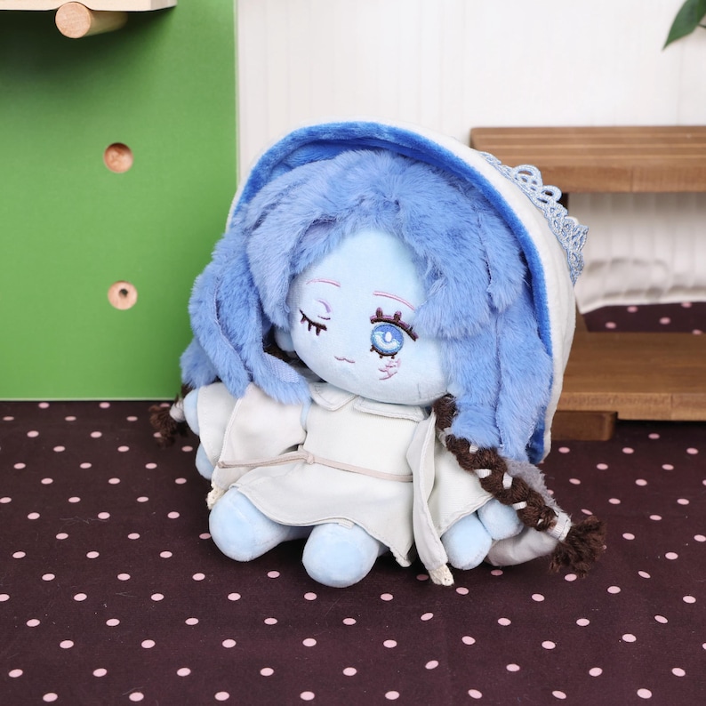 May include: A plush doll with blue hair and a white hooded cloak. The doll has closed eyes and a detailed face with embroidered features. The doll is sitting on a polka dot surface, with a green wall and wooden shelf in the background.