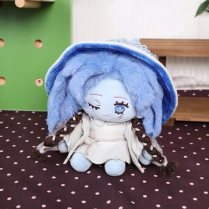 May include: A plush doll with blue fur and a matching hat. The doll has a white dress and brown braids. The doll has a detailed face with closed eyes and a single open eye. The doll is sitting on a brown surface with white polka dots.