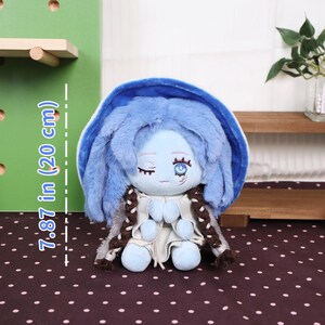 May include: A plush doll with blue hair and a matching hood, measuring 7.87 inches (20 cm) tall. The doll has a light blue face with closed eyes and a detailed eye design. It wears a white outfit with brown braids.