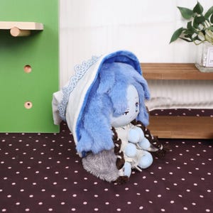 May include: A plush doll with blue fur and a white and blue lace-trimmed hood. The doll has a light blue face with embroidered features and brown braids. The doll is resting on a dark brown surface with white polka dots.