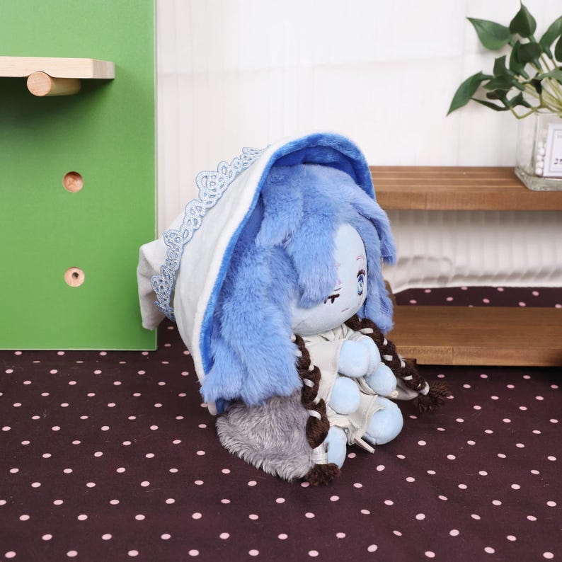 May include: A plush doll with blue fur and a white and blue lace-trimmed hood. The doll has a light blue face with embroidered features and brown braids. The doll is resting on a dark brown surface with white polka dots.