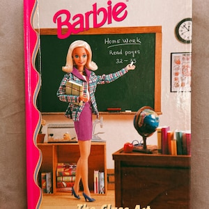 Peut inclure: Couverture de livre Barbie, intitulée "The Class Act", avec une poupée Barbie habillée en enseignante. La couverture a un dos rose et montre un tableau noir avec des instructions de devoirs. Un globe et des livres sont sur un bureau en bois.
