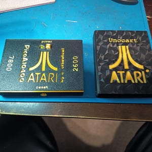 Peut inclure: Deux cartouches de jeu Atari noires. L'une porte l'inscription "Pico A10400" et "7800", avec le logo Atari en or. L'autre porte l'inscription "Unocart", avec le logo Atari en or.