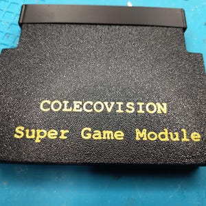 May include: A black textured ColecoVision Super Game Module cartridge with a smooth black top edge. The text COLECOVISION is printed in yellow capital letters above the yellow text Super Game Module. A small circular hole is visible on the left side.