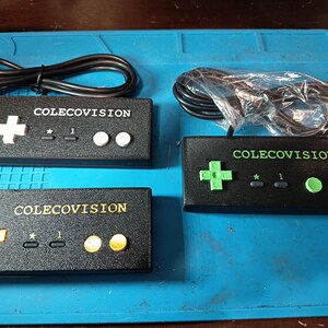 Peut inclure: Quatre manettes de jeu ColecoVision noires avec des croix directionnelles, des boutons et le texte "COLECOVISION". Les manettes ont des accents blancs, jaunes et verts. Des câbles sont attachés à certaines manettes, et l'une est dans un sac en plastique.