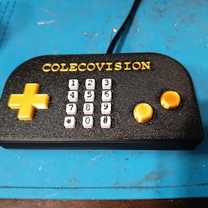 Peut inclure: Manette noire ColecoVision avec des boutons et une croix directionnelle dorés. La manette comporte un pavé numérique et le mot COLECOVISION en lettres dorées. Un câble noir s'étend du haut.