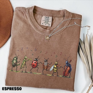 Può includere: Una t-shirt marrone a maniche corte con una grafica di sei insetti che suonano strumenti musicali, tra cui uno scarabeo alla batteria, una cavalletta alla tromba, una coccinella, un'ape al violino e uno scarabeo al violoncello.