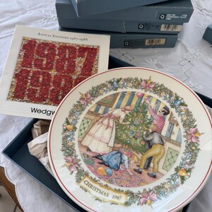 May include: A vintage Christmas plate from 1987, depicting a family decorating a Christmas tree. The plate is white with a detailed border and comes with its original box and a booklet. The text "Christmas 1987" is displayed on the plate.