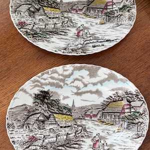 Può includere: Due piatti ovali in ceramica con una scena dettagliata di un paesaggio rurale. Il design include case con tetto di paglia, il campanile di una chiesa, alberi e figure che camminano lungo un sentiero, nei colori marrone, verde e rosso.