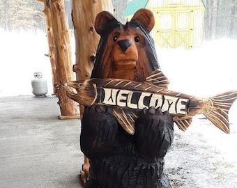 Puede incluir: Un oso de madera tallado sosteniendo un pez con la palabra "WELCOME" tallada en él. El oso está de pie sobre una base de madera.