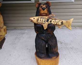 May include: A carved wooden bear holding a fish with the word "WELCOME" carved into it. The bear is brown and black and the fish is light brown. The bear is standing on a wooden base.