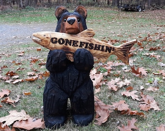 Puede incluir: Una talla de madera de un oso negro sosteniendo un pez de madera con el texto "GONE FISHIN'" tallado en él. La talla está sobre un lecho de hojas marrones.
