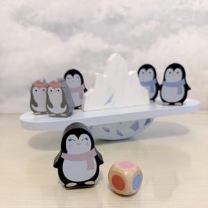 May include: A wooden penguin balancing game featuring a white iceberg, a dice, and several penguin figures with scarves and hats. The game is set on a light blue platform and base, creating a playful, wintery scene.