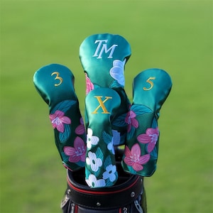 Könnte beinhalten: Ein Satz von vier teal Golfschlägerhauben mit floraler Stickerei. Die Hauben zeigen die Zahlen 3 und 5, den Buchstaben X und die Initialen TM in Weiß und Gold. Rosa und weiße Blumen verzieren die Hauben.