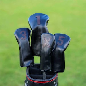 May include: A set of black golf club head covers with brown stitched numbers and an "X". The covers are in a golf bag with a red stripe. The background is a blurred green field.