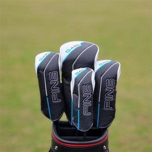 Puede incluir: Un conjunto de fundas para palos de golf en una bolsa. Las fundas son negras con detalles blancos y azules, y la marca "PING" es visible. El fondo es un campo verde borroso.