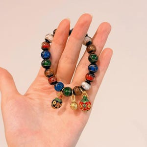 May include: A beaded bracelet featuring a variety of colorful, round beads in shades of red, blue, green, brown, and white. The bracelet has three decorative charms in gold and black with intricate designs.