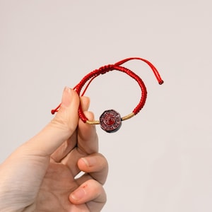 May include: Red braided bracelet with a central octagonal charm. The charm is dark red with a circular design. The bracelet is held against a plain background.