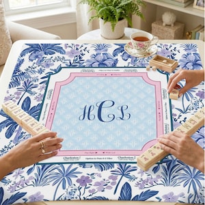 May include: A blue and white floral patterned Mahjong game table cover with a light blue center featuring the initials HCL. The cover has a pink border and is surrounded by hands holding Mahjong tiles. A cup of tea and a small wooden box with dice are also visible.