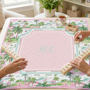 Puede incluir: Un tapete de juego de Mahjong rosa y turquesa con un diseño tropical que presenta palmeras y una escena de playa. El tapete tiene un área central rosa con un patrón de clave griega y las iniciales "JEC". Se ven fichas de Mahjong de madera.