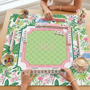 May include: A Mahjong game in progress on a floral patterned mat. The mat features a green center with a pink border and the initials "MCG". Pink Mahjong tiles are arranged on the mat, with a cup of coffee and a small vase of flowers nearby.