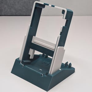 Peut inclure: Un support de téléphone en plastique teal et blanc. Le support a une base rectangulaire et une structure de support inclinée avec une fente pour tenir un téléphone. Le design comprend des lignes verticales et un accent blanc sur le bord supérieur.
