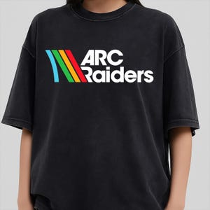 Può includere: T-shirt nero con la scritta bianca "ARC Raiders" e una grafica colorata a strisce blu, verdi, gialle e rosse. La maglietta ha maniche corte e una vestibilità ampia.