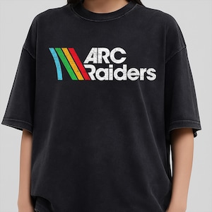 Può includere: T-shirt nero con la scritta "ARC Raiders" in bianco, accompagnata da un design a righe colorate in blu, verde, giallo e rosso. La maglietta ha un aspetto vintage e usurato.