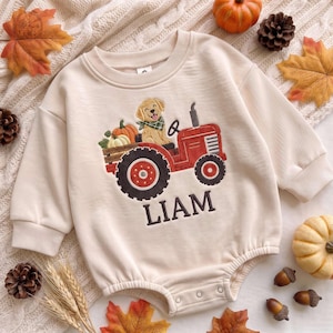 May include: A cream-colored long-sleeve baby romper featuring an embroidered red tractor with a golden retriever puppy wearing a green plaid bandana, pumpkins in the back, and the name LIAM embroidered in dark brown lettering below the tractor.
