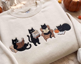 May include: A cream-colored crewneck sweatshirt featuring an embroidered design of four black cats. One cat lies on a pillow reading, one holds a stack of books, one sits in a brown armchair reading, and one sleeps on an open book next to a pumpkin.