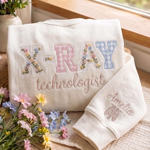 May include: A cream-colored crewneck sweatshirt featuring the embroidered text X-RAY in a mix of floral and gingham patterns, with the word technologist below. The sleeve displays the name Amelia above an embroidered rib cage illustration.